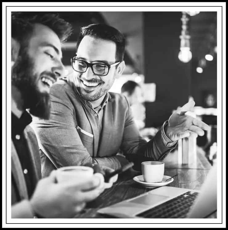 2 men laughing holding coffee mugs and sitting by a computer